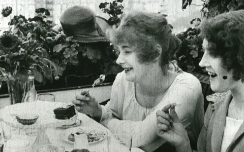 Två kvinnor, en av dem Greta Gustafsson, skrattar vid ett bord med tårta och blommor.