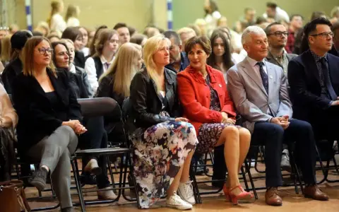 Uczestnicy siedzą na krzesłach, słuchając uważnie. Wśród nich widać osoby, które mogą być na radzie pedagogicznej, w tym kobiety w eleganckich strojach i mężczyzn w garniturach.