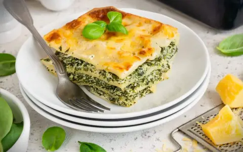 Ein Stück Spinatlasagne mit Käse überbacken, garniert mit Basilikum, auf einem Teller mit einer Gabel.