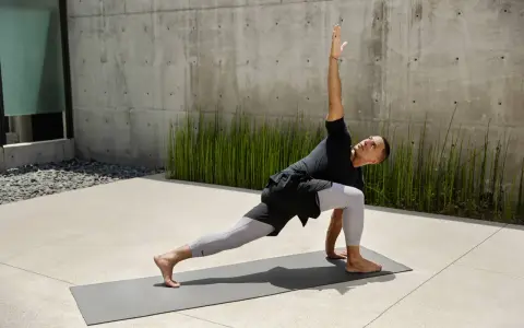 Mann in Yoga-Pose auf Matte vor Betonwand und grünen Pflanzen.