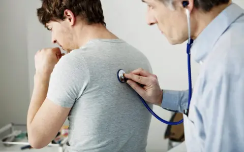 Arzt untersucht Patienten mit Stethoskop. Der Patient hustet, was auf Kurzatmigkeit hindeuten könnte.