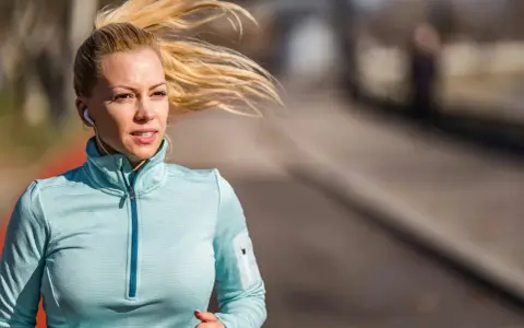 Kvinna med blont hår i hästsvans springer utomhus, lyssnar på musik med hörlurar. Hon är fokuserad på att springa 5 km tid.