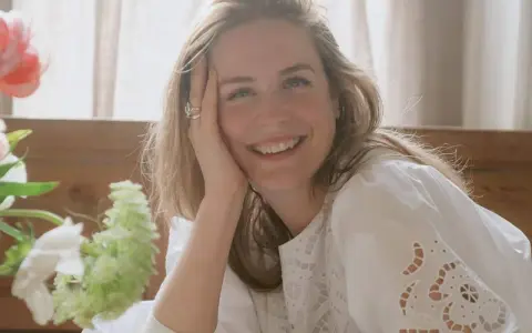 Glad kvinna med blont hår ler mot kameran, med handen mot kinden. En bukett blommor syns till vänster. Kanske ett foto från sofia wood blogg.