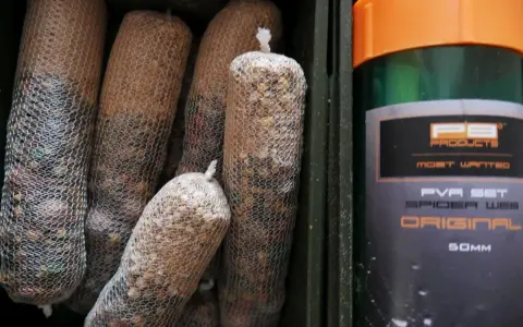 Wędkarskie stiki w siatkach PVA, gotowe do użycia. Obok zielony pojemnik z pomarańczową nakrętką.