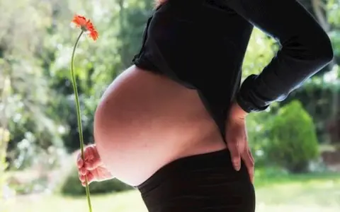 Brzuszek w 3 miesiącu ciąży, delikatnie podtrzymywany dłonią, z kwiatkiem w tle.