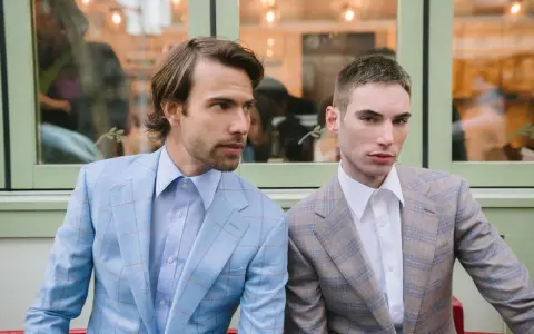 Two men in plaid suits with wide notch lapels sit side-by-side. The man on the left wears a light blue suit, the man on the right a tan suit.