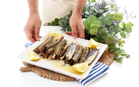 Un plato blanco con navajas a la plancha, limón y perejil. Unas manos sostienen el plato, decorado con eucalipto y flores moradas.