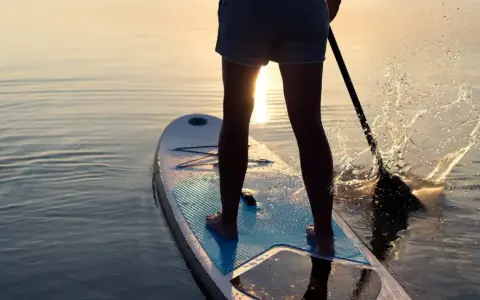 Person lernt SUP auf ruhigem Wasser bei Sonnenuntergang. Das Paddel spritzt Wasser auf.