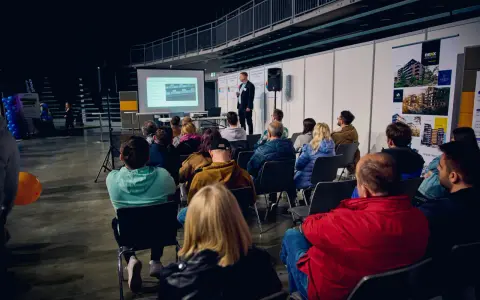 Prezentacja na targach budowlanych. Mężczyzna przemawia do zgromadzonej publiczności, która siedzi na krzesłach.