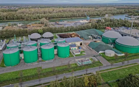 Grüne Biogas-Behälter und große Gasspeicher prägen diese Anlage, die die **Biogas Zukunft** symbolisiert. Im Hintergrund ein Fluss und bewaldete Ufer.