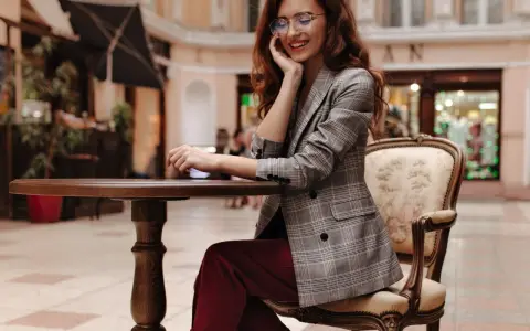 Elegancka kobieta w okularach i marynarce w kratę, prezentująca stylizacje z bordowymi spodniami, siedzi przy stoliku w kawiarni.
