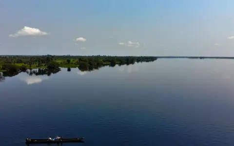 Szeroka, spokojna rzeka Kongo, odbijająca niebo i bujną zieleń brzegów. Na wodzie płynie łódź z kilkoma osobami.