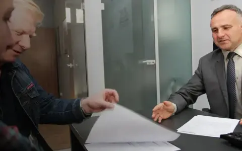 Dwóch mężczyzn przy biurku przegląda dokumenty, jeden w garniturze, drugi w dżinsowej kurtce.