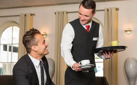 Un serveur souriant présente une tasse de café à un client, illustrant le protocole de service à table.
