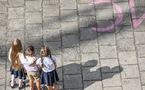 Trójka dzieci w szkolnych strojach patrzy na napis "szkoła społeczna co to" narysowany kredą.