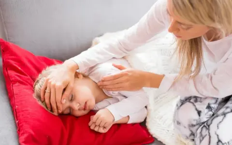 Matka troskliwie opiekuje się dzieckiem, które leży na czerwonej poduszce. Dziecko ma zamknięte oczy, a matka delikatnie dotyka jego czoła, sprawdzając, czy nie ma gorączki. To obrazek pokazujący troskę o dziecko podczas jelitówki.
