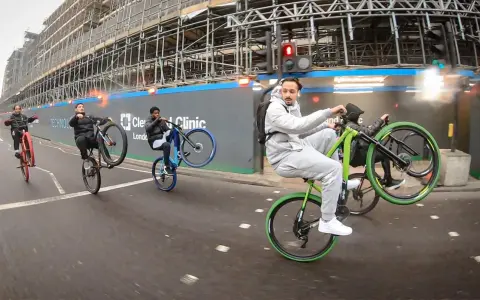 Młodzi mężczyźni wykonują **jazdę na jednym kole** na rowerach, mijając plac budowy. Jeden z nich, w szarym dresie, robi to na zielonym rowerze.