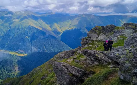 Dwie osoby wędrują po skalistym szlaku w górach Fogaraskich. W tle rozciągają się zielone pasma górskie i lasy.