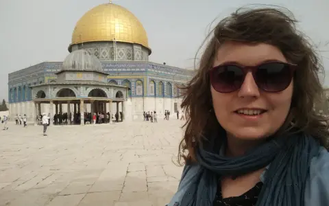 Uśmiechnięta kobieta w okularach przeciwsłonecznych robi selfie na tle Kopuły na Skale. Wokół widać turystów, co sugeruje, że w Jerozolimie jest bezpiecznie.