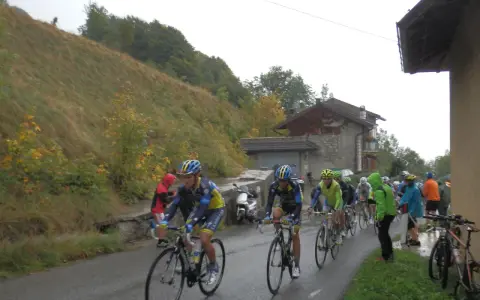 Ciclisti in gara su una strada bagnata, con una **forcella alta** che indica la salita. Il paesaggio è autunnale, con alberi e un pendio erboso.