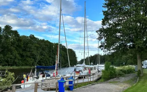 Segelbåtar vid kajen på Dalbergså camping. Grönskande träd och en blå himmel med moln.