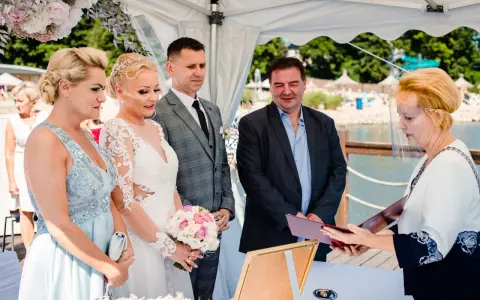 Ceremonia ślubna nad jeziorem, panna młoda w białej sukni, pan młody w garniturze, urzędniczka w przyłbicy.