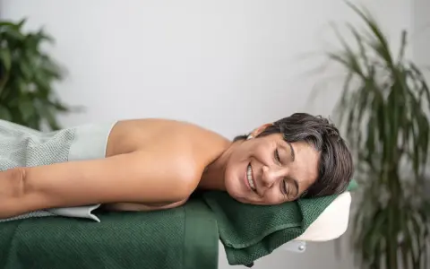 En kvinna ler och njuter av att vila på en massagebänk, insvept i en ljusgrön handduk. Gröna växter i bakgrunden bidrar till en lugn atmosfär.