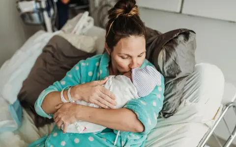 Mama tuli dziecko po cesarce, czule całując je w czapeczkę. Laktacja po cesarce może być wyzwaniem, ale miłość jest silniejsza.