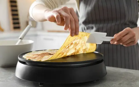 Une personne utilise une spatule pour soulever une crêpe dorée de la plaque chauffante. Astuce pour comment nettoyer une crêpière électrique.