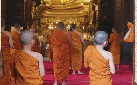 A group of Buddhist monks, another word for ascetics, walk towards a large golden Buddha statue in a temple.