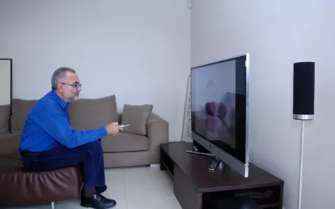 Starszy mężczyzna w okularach siedzi na kanapie i ogląda telewizję, trzymając pilota.