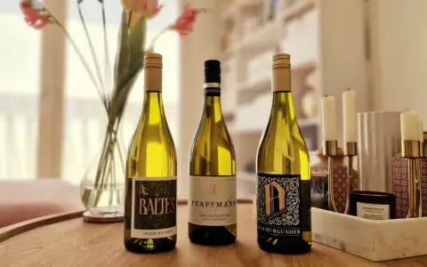 Drei Flaschen Grauburgunder Wein stehen auf einem Holztisch, daneben Blumen und Kerzen.