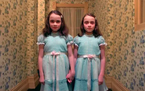 Dos niñas idénticas, vestidas con trajes azules, se toman de la mano en un pasillo con papel tapiz floral. Son las gemelas de "Bso El Resplandor".