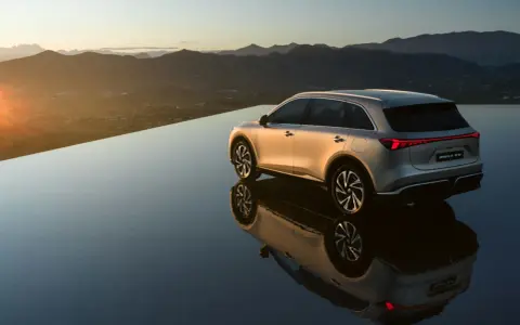 El MG6 EV plateado se refleja en una superficie de agua infinita al atardecer, con montañas al fondo.