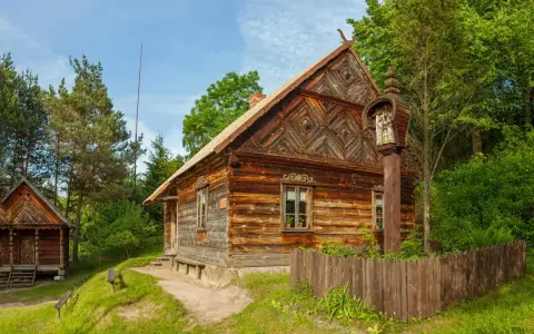 Drewniane chaty i kapliczka na słupie w jednym z **skansenów w Polsce**. W tle las i błękitne niebo.