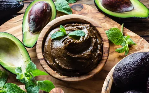 Crema di avocado vellutata in ciotola di legno, circondata da avocado freschi e foglie di menta.