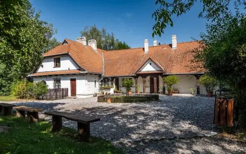 Urokliwy dom z czerwonym dachem w Bronowicach Małych, otoczony zielenią. Widać drewniane ławki i kamienny dziedziniec.