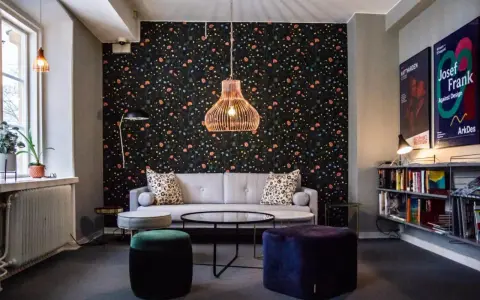 En mysig lounge med blommig tapet, en grå soffa, färgglada sittpuffar och en hylla full med böcker. En affisch för "Josef Frank - Against Design" hänger på väggen.