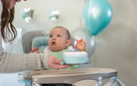 Mama podaje tort z jedną świeczką na roczek maluszkowi. Balony i dekoracje tworzą radosną atmosferę.