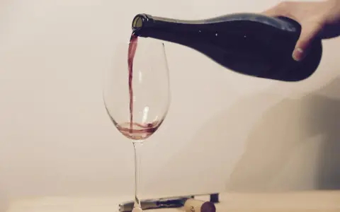 Ein Weinglas wird mit Rotwein gefüllt. Eine Hand hält die dunkle Flasche, während der Wein in einem dünnen Strahl ins Glas fließt. Ein Korkenzieher und ein Korken liegen daneben.