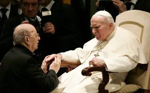 Papież Jan Paweł II w białej szacie, klęczący mężczyzna w czerni. W tle widać fotografów. To obraz duchowej siły, jakiej doświadczają legiony Chrystusa.