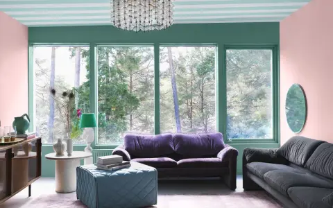 Ett vardagsrum med art deco inredning, mjuklila soffa, mörkgrå soffa, turkos pall och en stor fönsterutsikt.