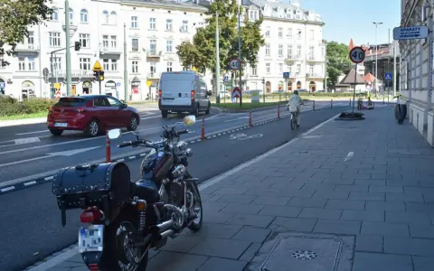 Motocykl zaparkowany na chodniku, obok drogi z wyznaczonym pasem rowerowym. W tle budynki i ruch uliczny, w tym czerwony samochód i biały van.