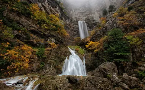 Cascada en un cañón rocoso, con árboles otoñales y niebla. El agua cae como un zafiro lagunazo, creando un espectáculo natural.
