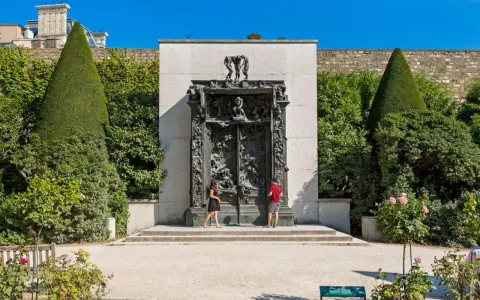 La Porte de l'Enfer, une œuvre monumentale de Rodin, se dresse majestueusement, encadrée par des arbres taillés et un mur de pierre. Deux visiteurs contemplent cette sculpture complexe.