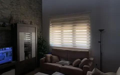 Salón acogedor con estores enrollables noche y día que filtran la luz, sofá, mueble y televisión.