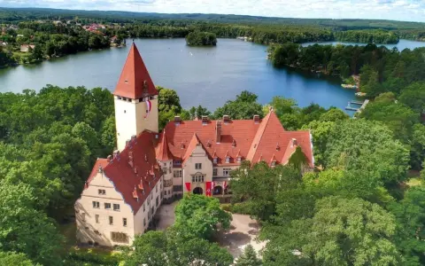 Zamek w Lubniewicach otoczony drzewami, nad jeziorem.