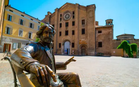 Statua bronzea in una piazza di una città dell'Emilia Romagna, con un'antica chiesa in mattoni sullo sfondo.