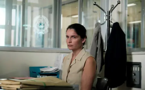 Una donna con un'espressione pensierosa siede in un ufficio disordinato, circondata da fascicoli. Sembra l'inizio di una storia nera.