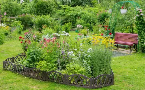 Un exuberante macizo de flores de verano, con rosas rojas, margaritas blancas y flores amarillas vibrantes, bordeado por una valla decorativa.